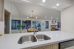 A modern kitchen with a double sink, a dining area with a table and chairs, and a view of a pool through large windows.