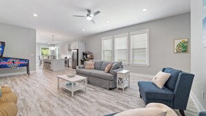 Ein modernes Wohnzimmer mit einem grauen Sofa, einem blauen Sessel und einem Couchtisch. Im Hintergrund befindet sich eine Küche mit einem Kühlschrank und einem Essbereich.