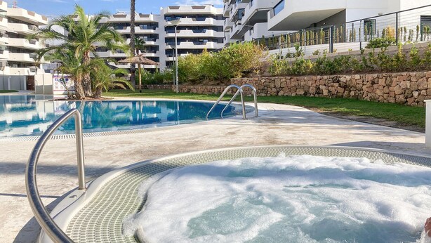 En swimmingpool med ett bubbelbad, omgiven av en stenmur och grönska, med moderna byggnader i bakgrunden.