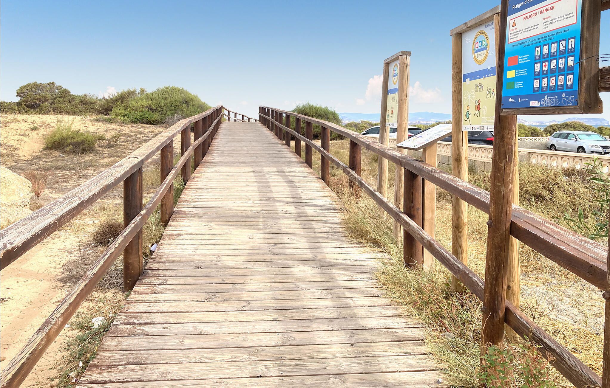 Ein hölzerner Steg führt zu einem Strandbereich, rechts ist ein Schild zu sehen.