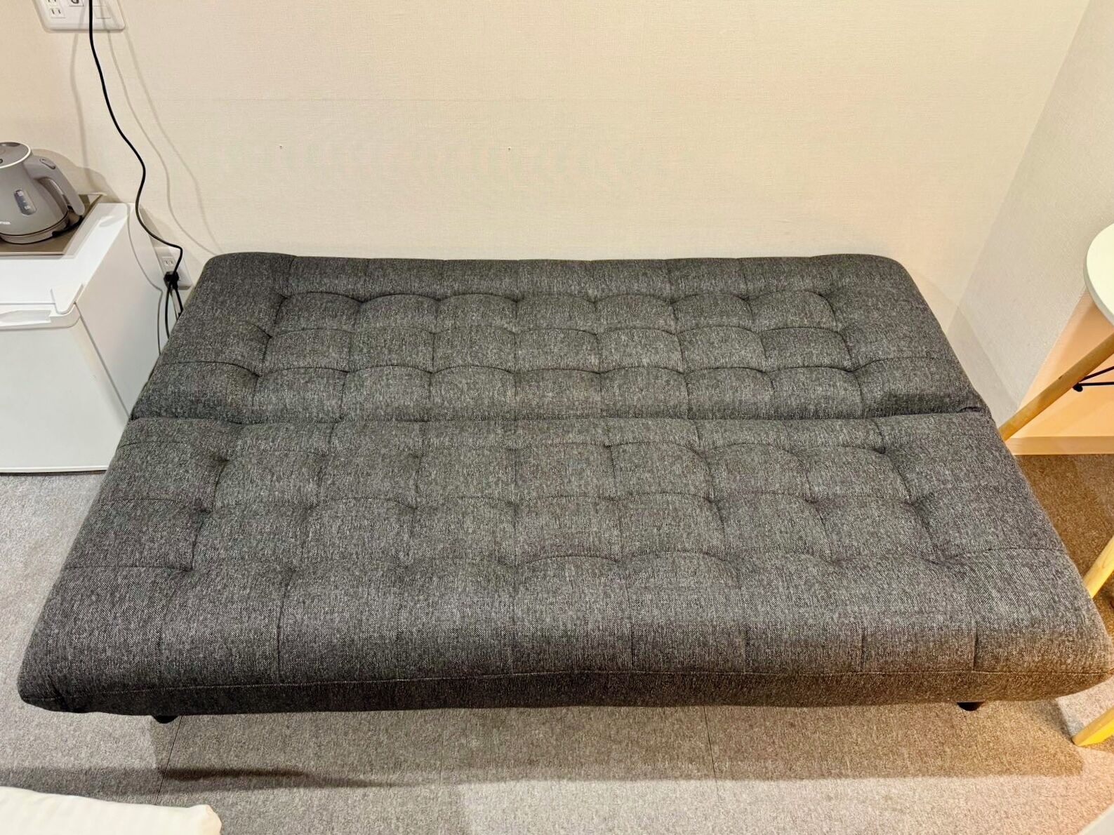Eine graue, gepolsterte Bank mit einer Noppenstruktur, platziert vor einer schlichten Wand mit einer Steckdose und einem kleinen Tisch links daneben.