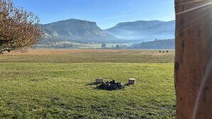 Blick durch eine Holzkonstruktion auf ein grasbewachsenes Feld, einen Baum und Berge im Hintergrund.