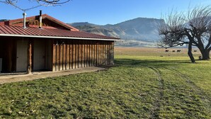Ein rustikaler Schuppen mit rotem Dach, Holzfassade und Betonfundament. Im Hintergrund erheben sich Berge und ein klarer, blauer Himmel.