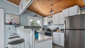 A kitchen with white cabinets, a stainless steel refrigerator, a stove, and a microwave. There are bar stools and a window above the sink.