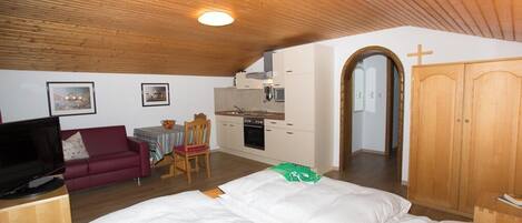 A bedroom with a wooden ceiling, a bed, a television, a red sofa, a dining table with chairs, and a kitchen area.