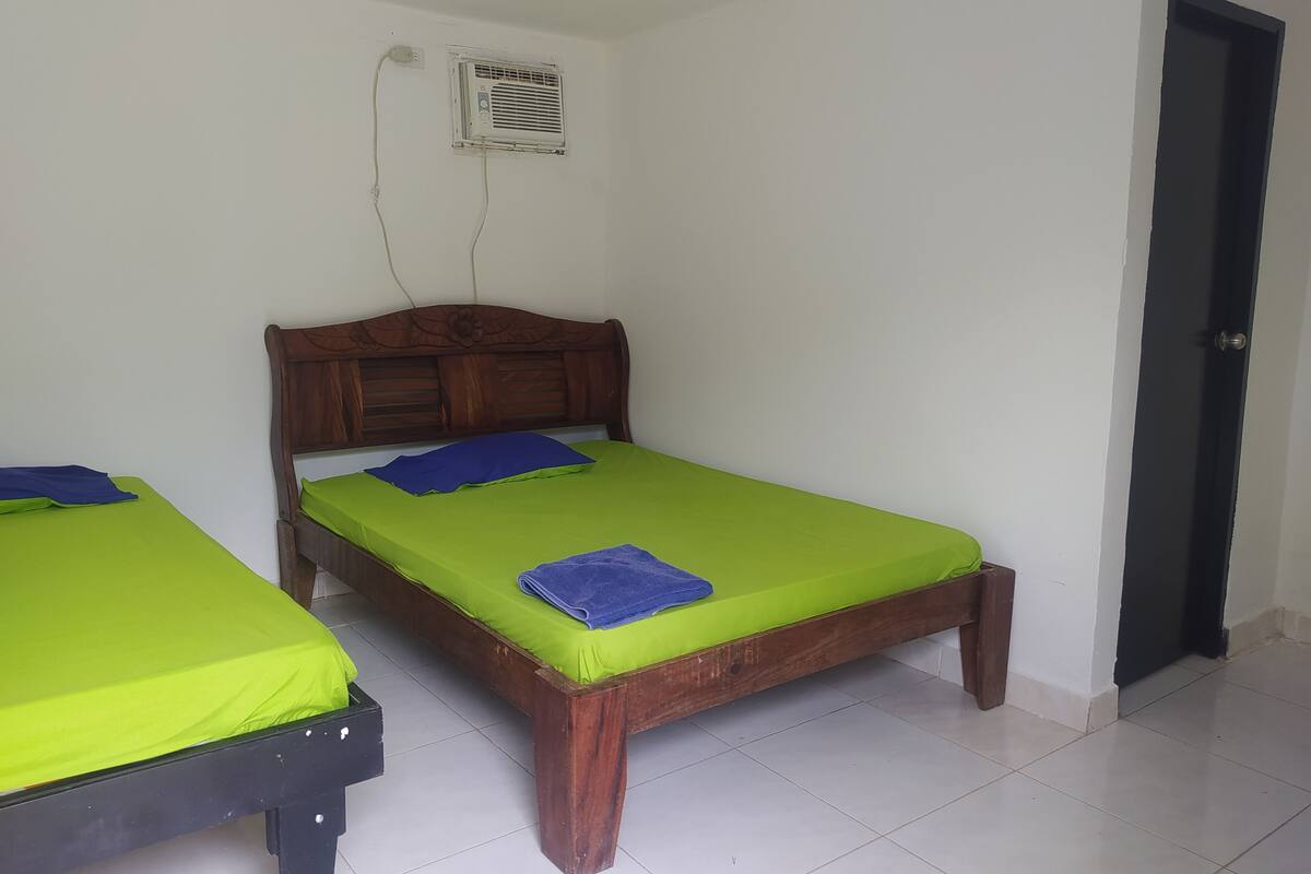 Une chambre avec deux lits en bois, chacun doté d’un matelas vert et d’un oreiller bleu. Un climatiseur mural est fixé au mur au-dessus des lits.