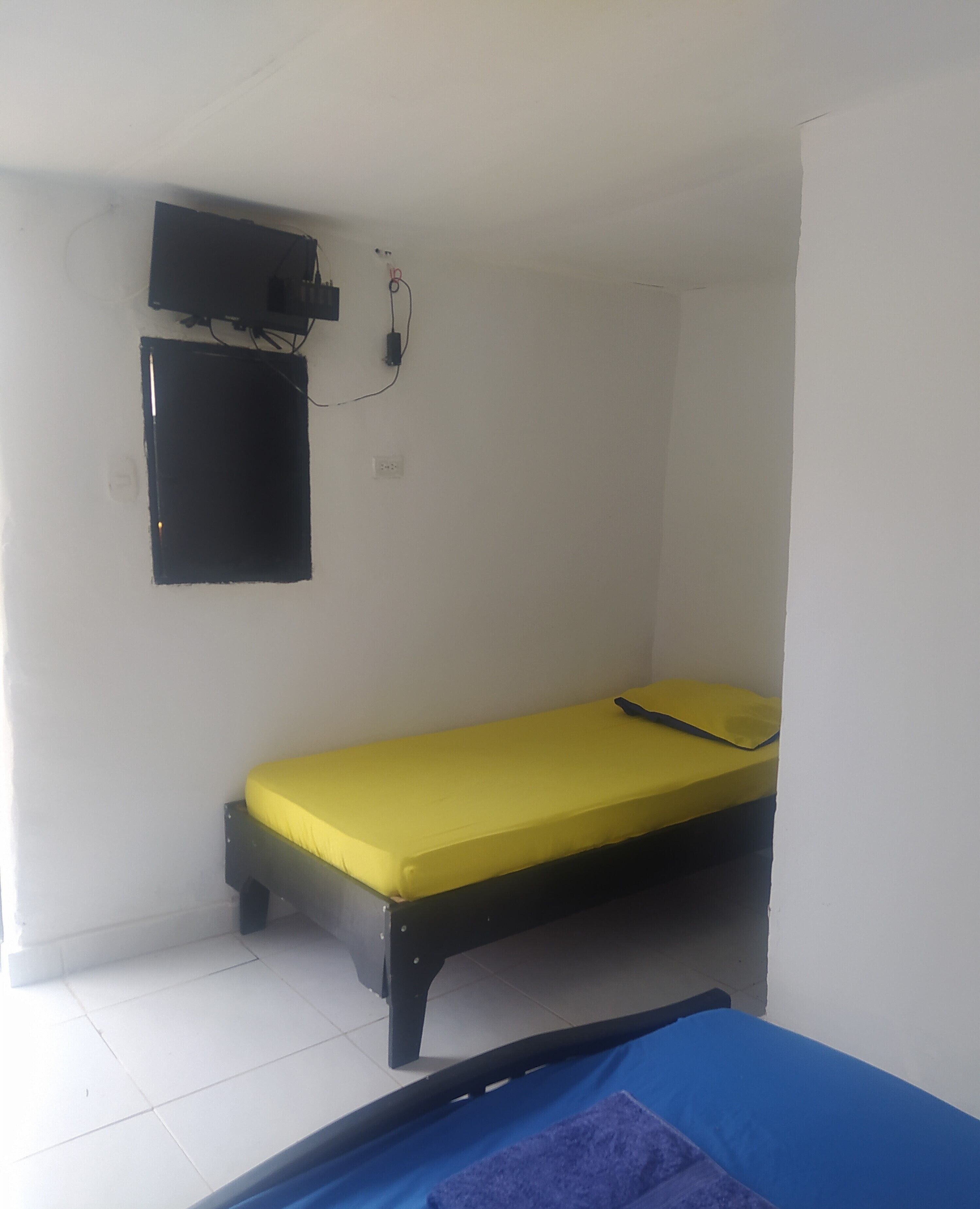 A small, single-bed room with a yellow mattress, a black headboard, a wall-mounted TV, and a power outlet.