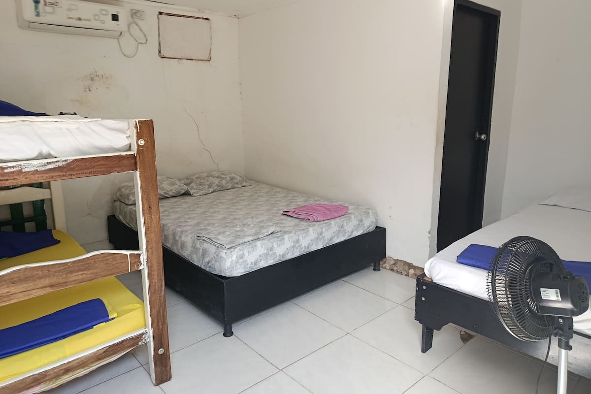 Une petite chambre simple, équipée d’un lit superposé, d’un lit simple et d’un ventilateur sur pied.