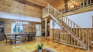 A spacious living area with a wooden staircase leading to an upper level, a dining area with a large table and chairs, and a cozy fireplace. - Sugar Grove Log Cabin | HOT TUB + Fire Pit! (Huntingdon)