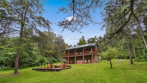 A rustic cabin with a deck, surrounded by lush greenery and tall trees. - Sugar Grove Log Cabin | HOT TUB + Fire Pit! (Huntingdon)