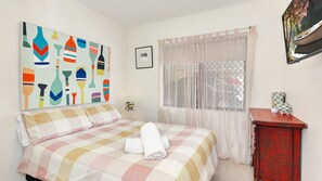 A bedroom with a bed, a window with sheer curtains, a painting on the wall, and a small wooden cabinet. - Ocean Sands 1 - 2 Bedroom Unit just a stroll to the beach (Sawtell)