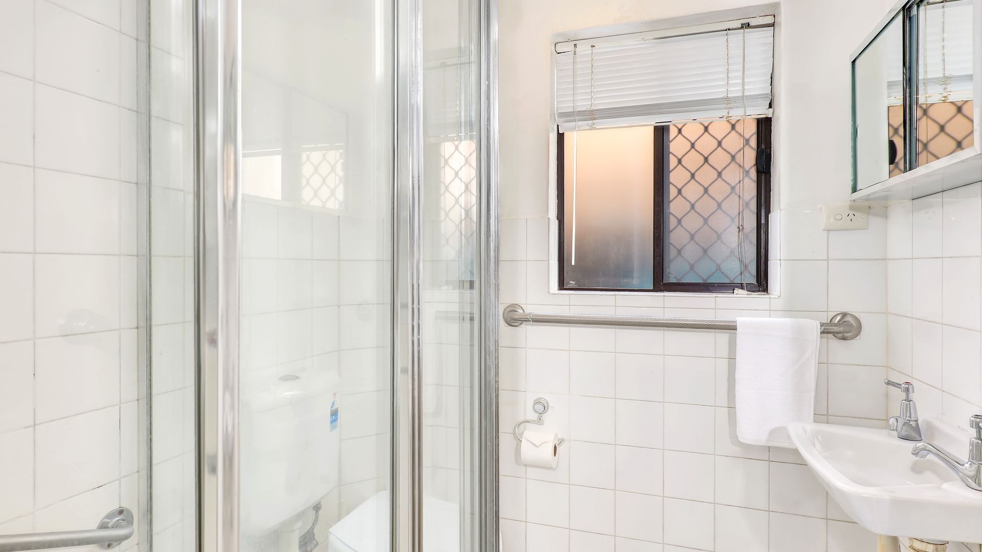 Un baño moderno con cabina de ducha de vidrio, un lavabo blanco y una ventana con persianas.
