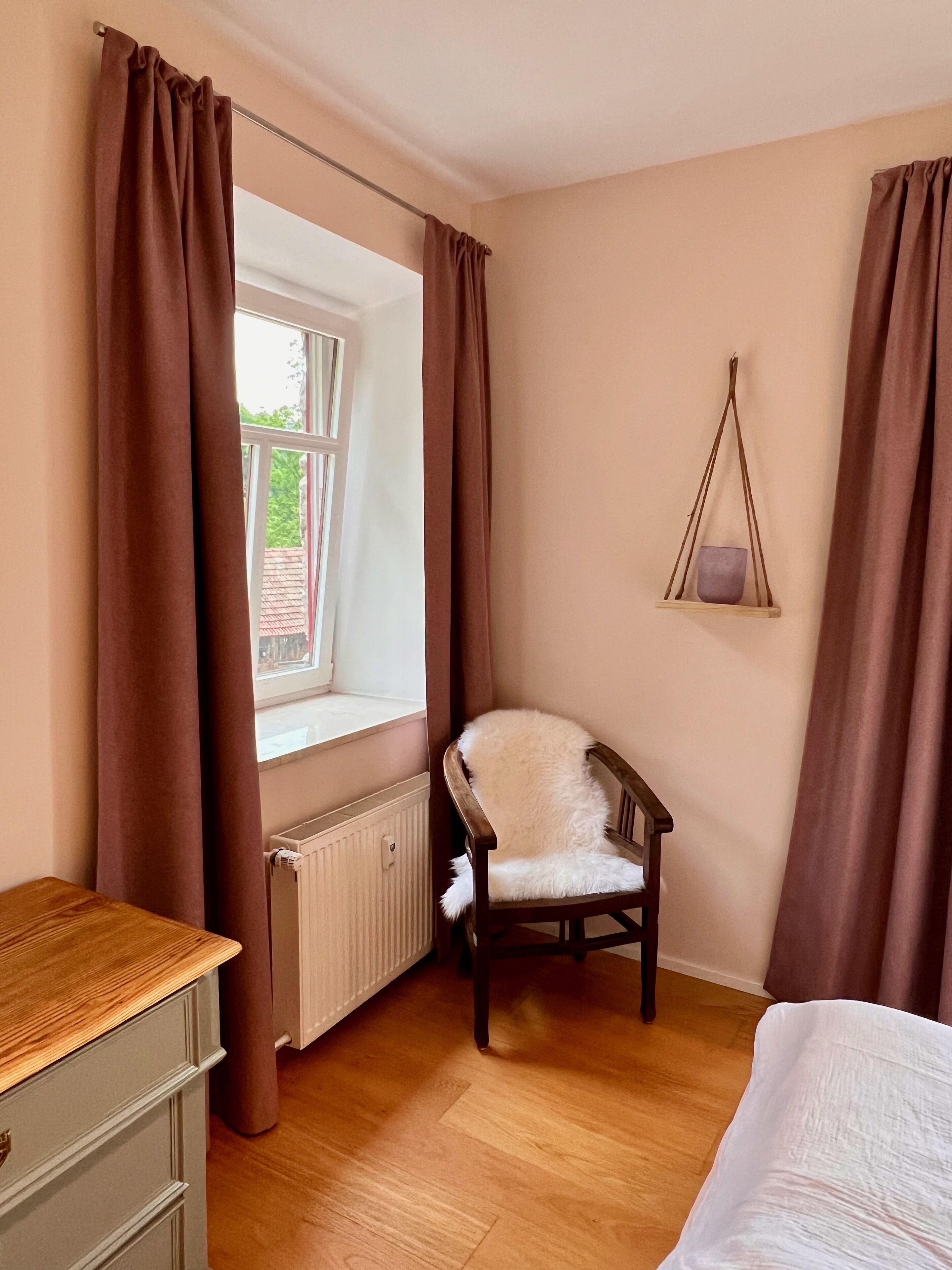 Un dormitorio con cómoda de madera, una silla, una ventana con cortinas y una planta colgante.