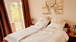 Ein großes Bett mit weißer Bettwäsche, zwei Nachttische mit Lampen, ein Fenster mit Vorhängen und ein Wandbild.