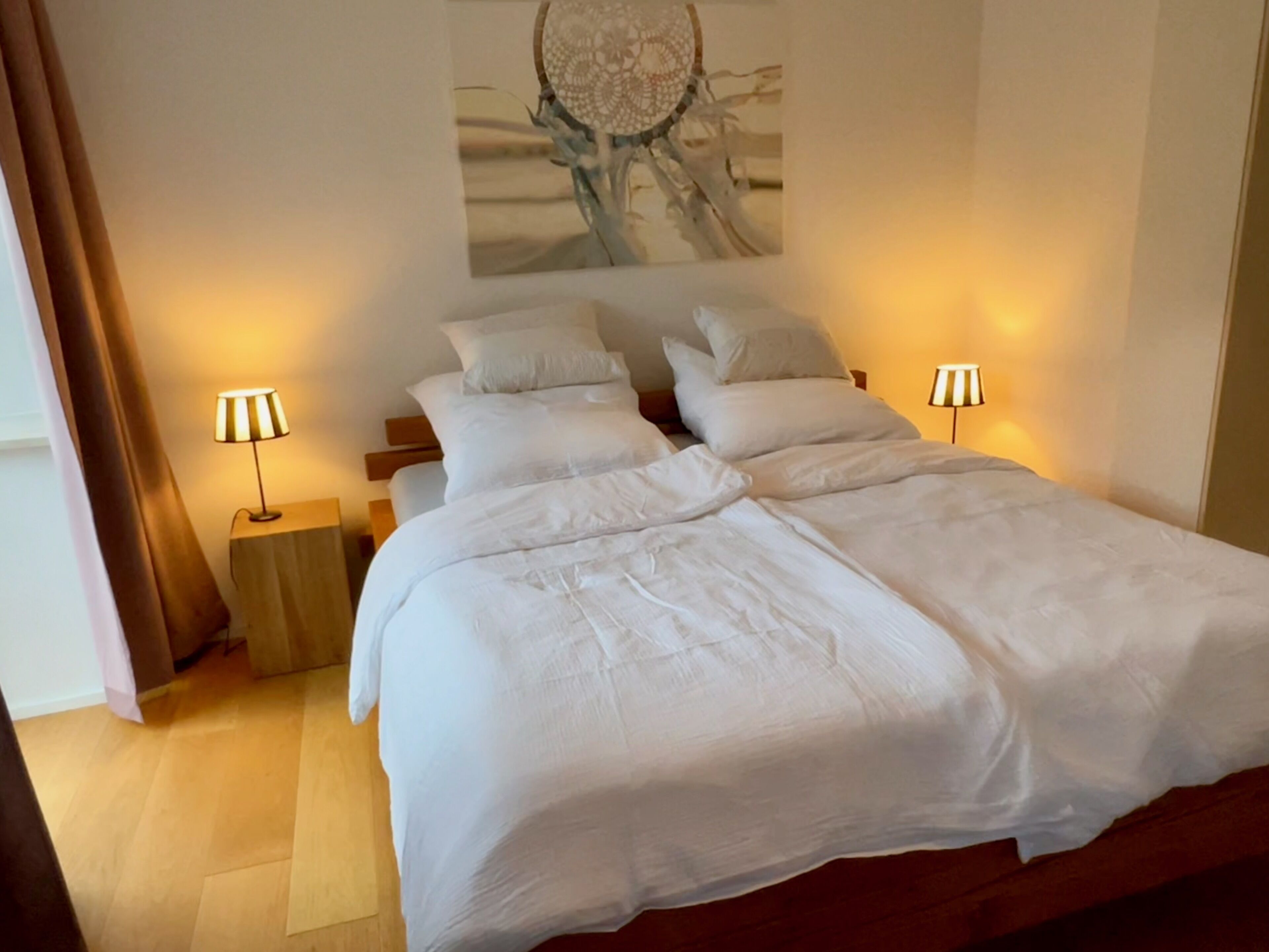 Ein ordentlich bezogenes Bett mit weißen Leinen, zwei Nachttischlampen und einer Traumfänger-Wandkunst.