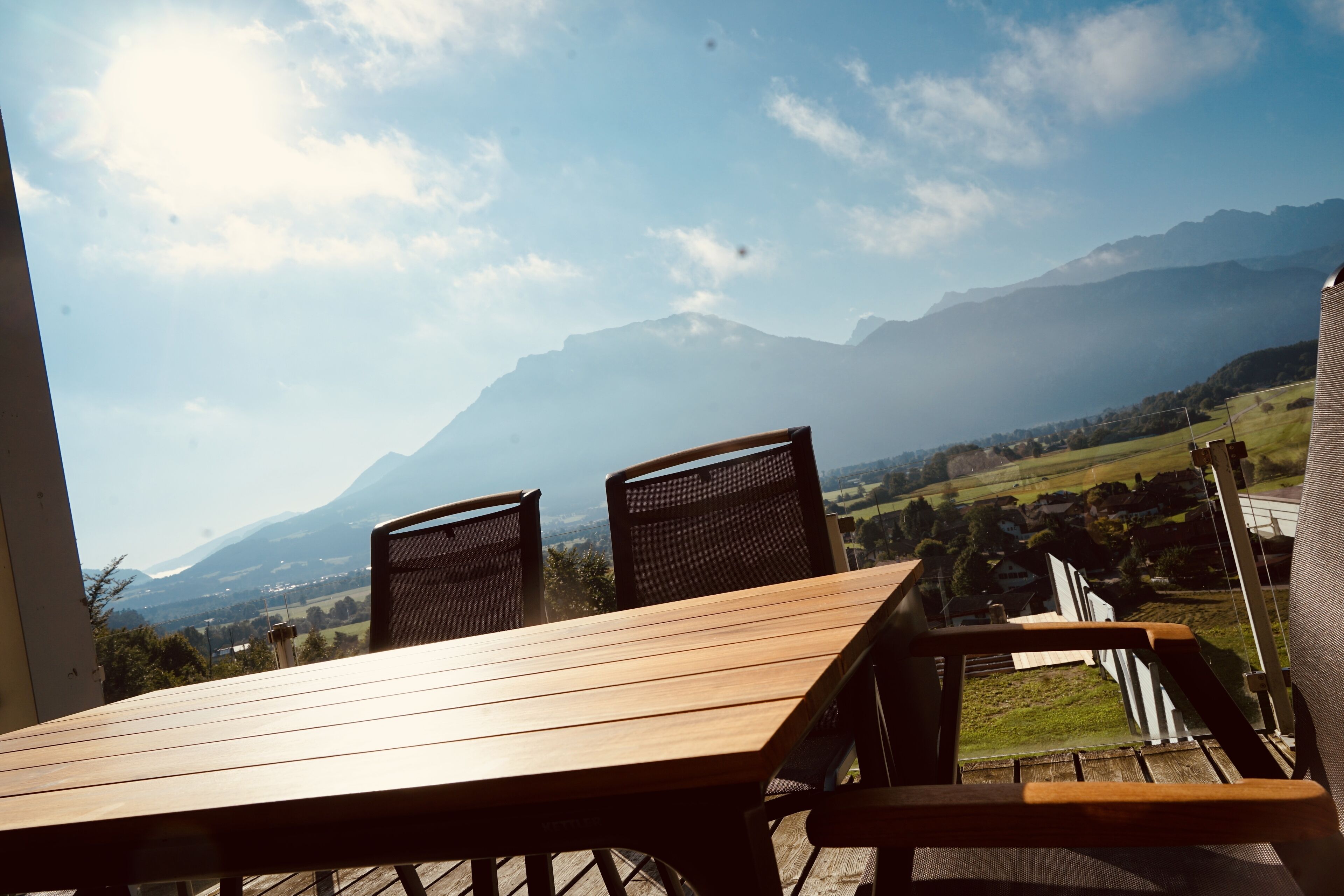 Ein Holztisch und Stühle auf einer Terrassenförmigen Außensitzgelegenheit mit Blick auf Berge und ein Dorf weiter unten.