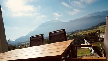 Ein Holztisch und Stühle auf einer Terrassenförmigen Außensitzgelegenheit mit Blick auf Berge und ein Dorf weiter unten.
