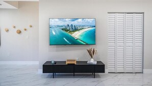 A modern living room with a large flat-screen TV mounted on the wall, a black console table with a book and a vase, and a white door with horizontal blinds. - Miami 4BR Family Home | Patio + Parking (Miami)