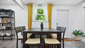 A dining area with a dark wooden table and chairs, a potted plant, and a window with yellow curtains.