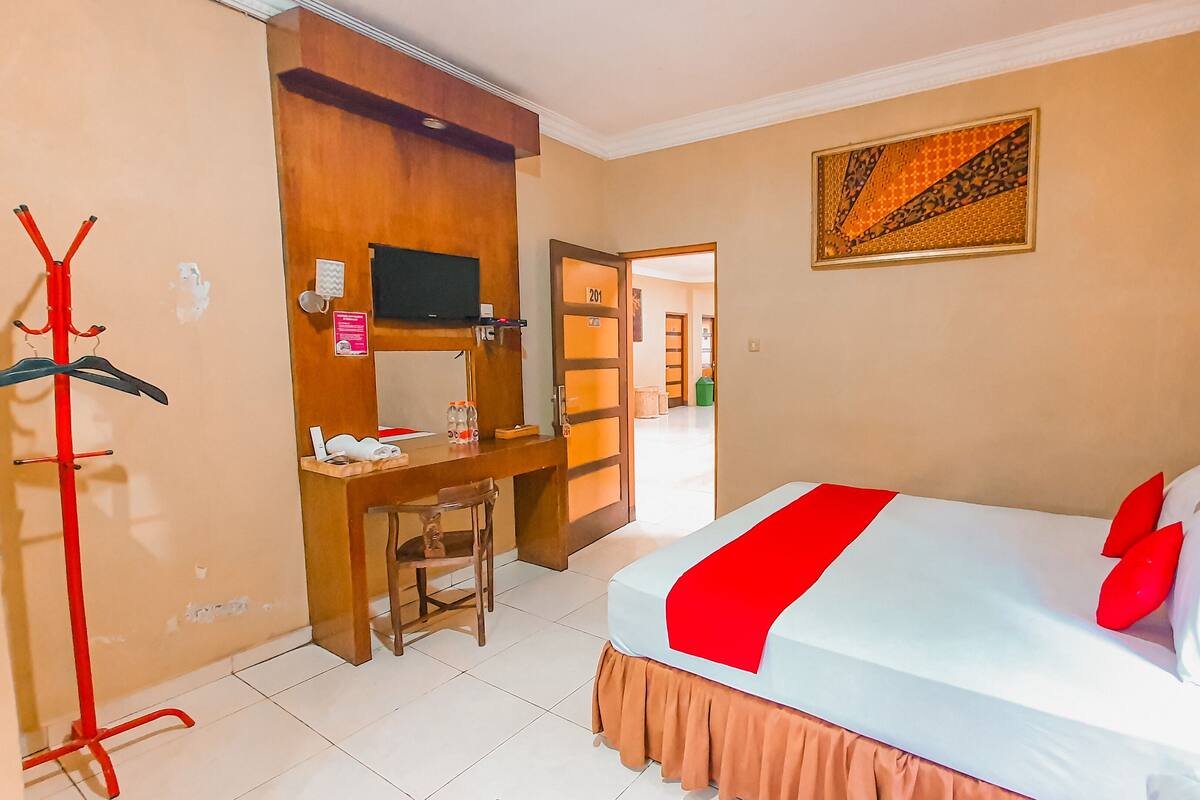 Habitación de hotel con cama, escritorio, silla, perchero y televisión.
