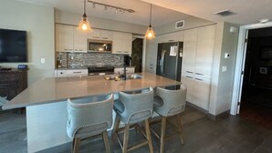 Une cuisine moderne dotée d’un grand îlot central, de tabourets de bar, de luminaires suspendus et d’appareils électroménagers en acier inoxydable.