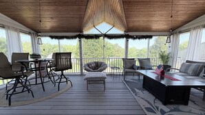 A covered outdoor patio with a wooden ceiling, a round table, chairs, a sofa, and a view of trees.