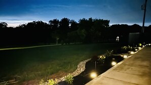 A nighttime view of a landscaped area with a pathway, small plants, and a grassy field.