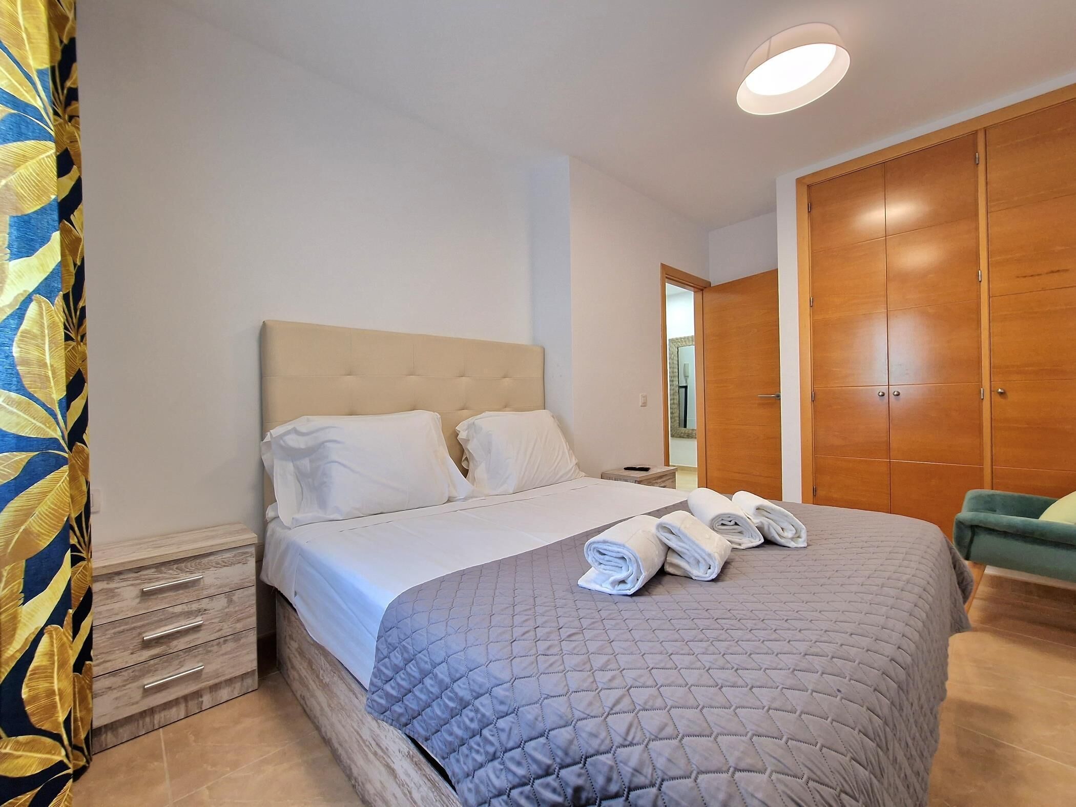 A neatly made bed with a grey quilt, a wooden wardrobe, and a green armchair.