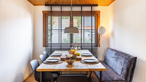 Un coin repas avec une table en bois, des chaises noires, un banc et une grande fenêtre équipée de stores.