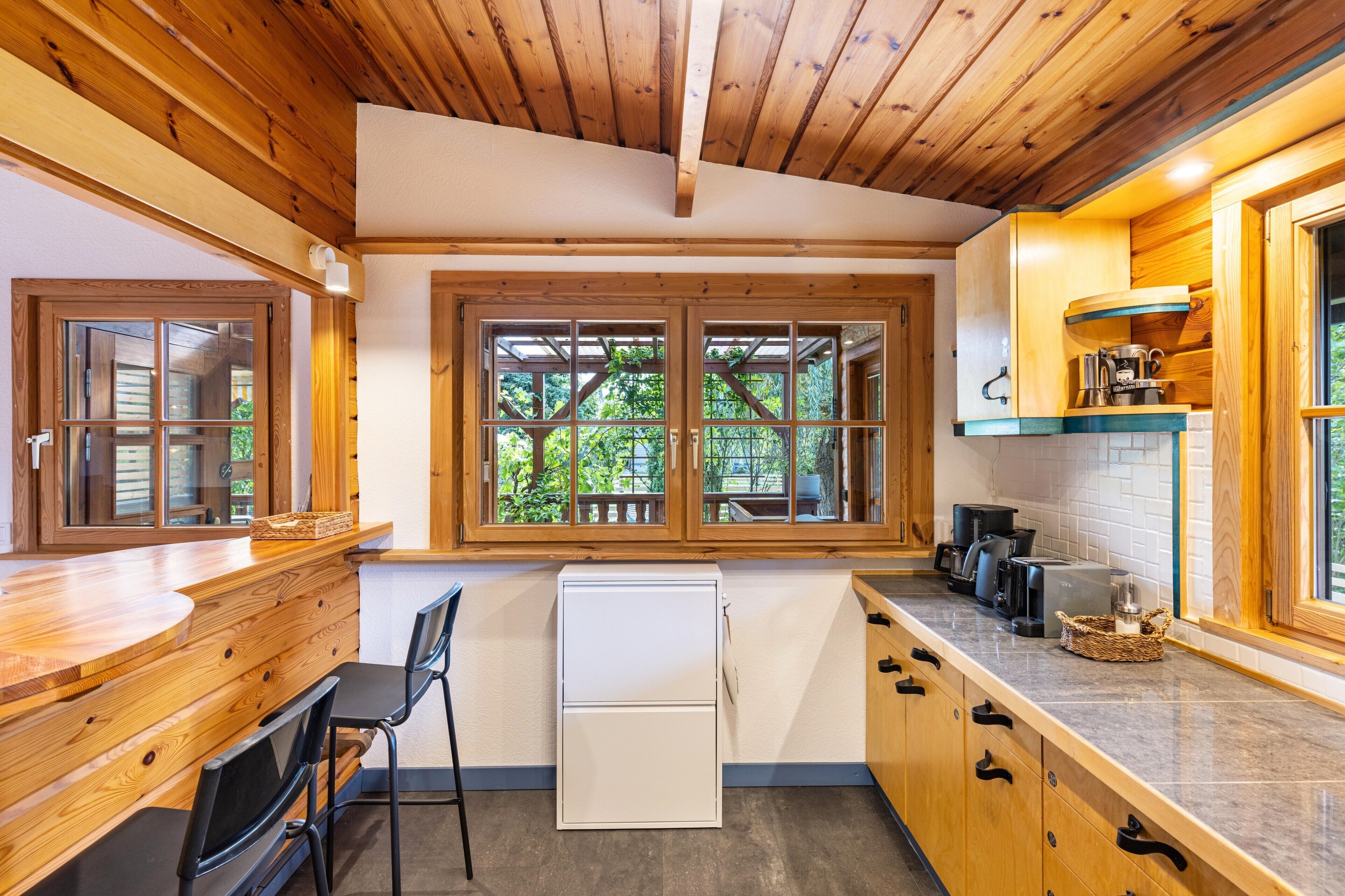 A compact kitchen with wooden cabinets, a built-in refrigerator, and a window overlooking greenery.