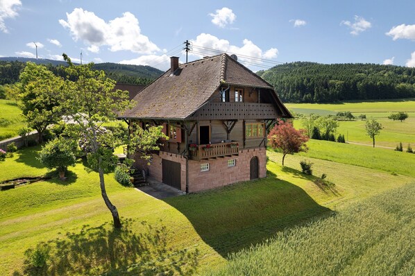Une maison traditionnelle en bois, dotée d’un balcon, entourée de champs verdoyants et bordée, à l’arrière-plan, d’une colline boisée.
