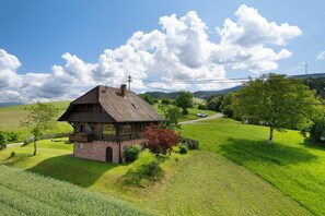 A traditional house with a sloped roof, surrounded by lush green fields and a clear sky.