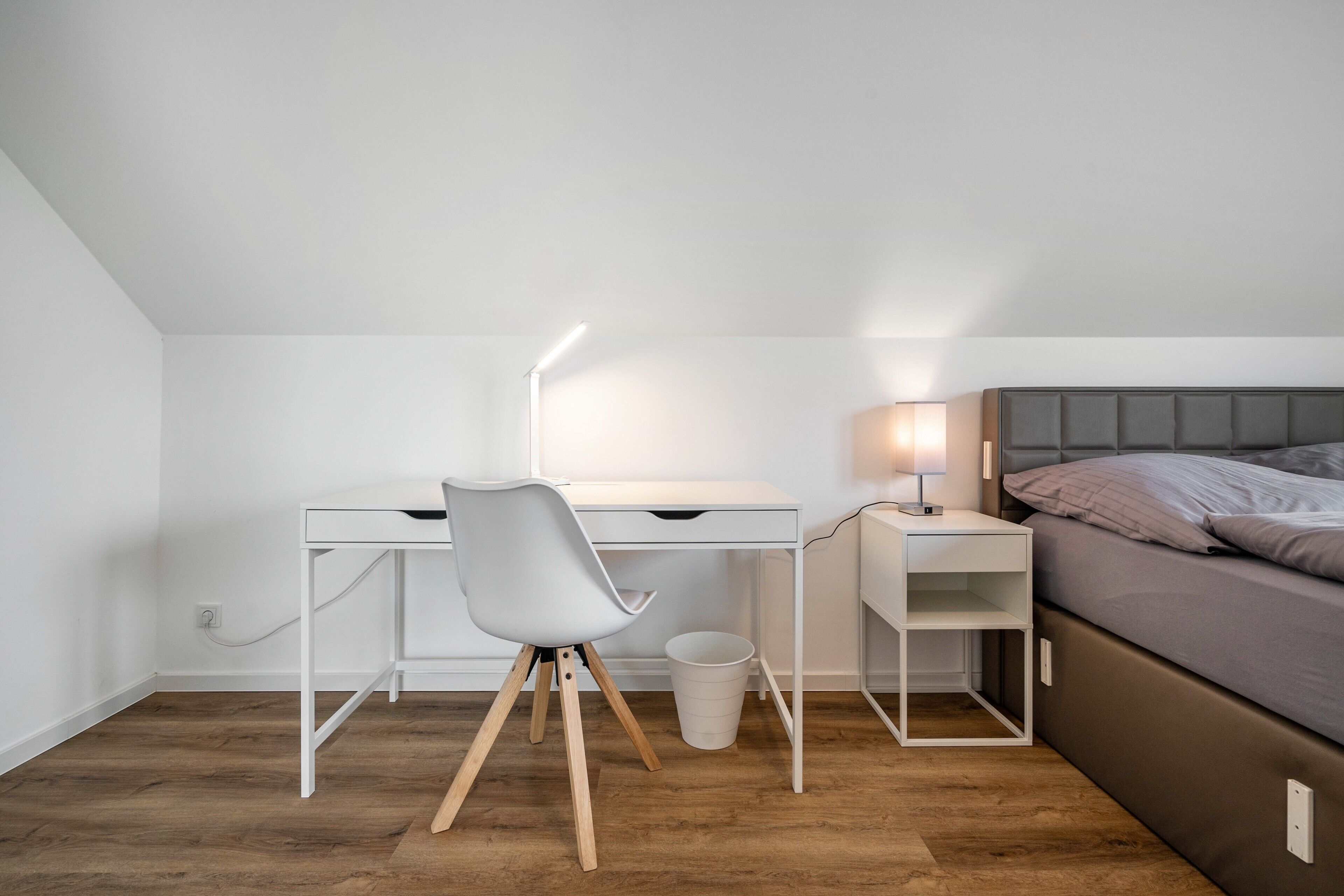A bedroom with a bed, a desk with a chair, a bedside table with a lamp, and a white wall.