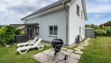 Ein Haus mit Terrasse, Grill und zwei weißen Liegestühlen.