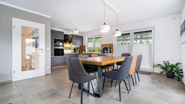 Una cocina moderna con comedor, que incluye una mesa de madera, sillas tapizadas en gris, iluminación colgante y un amplio ventanal con persianas.