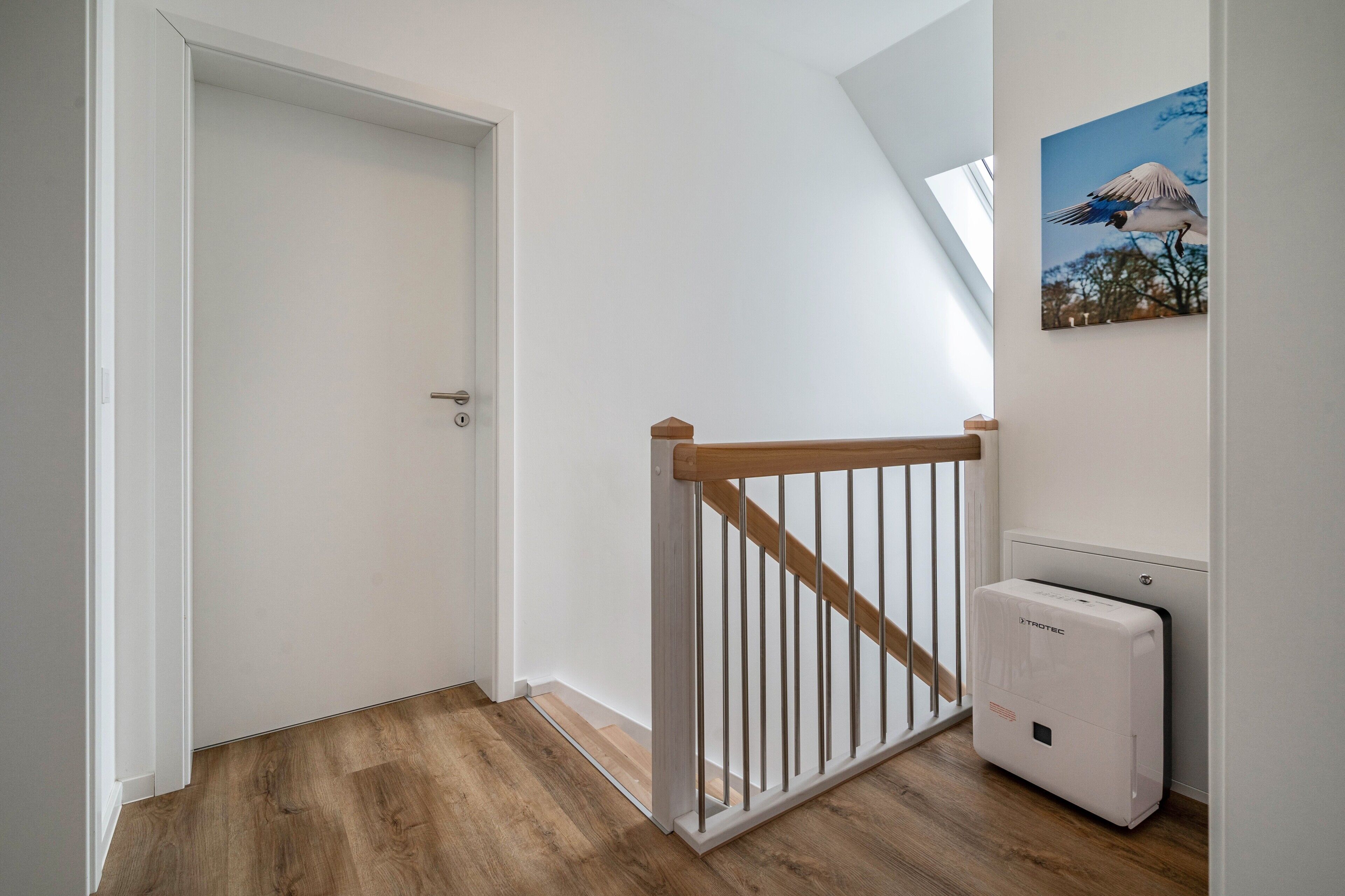 A room with a wooden floor, a white door, a white railing, and a white device on the wall.