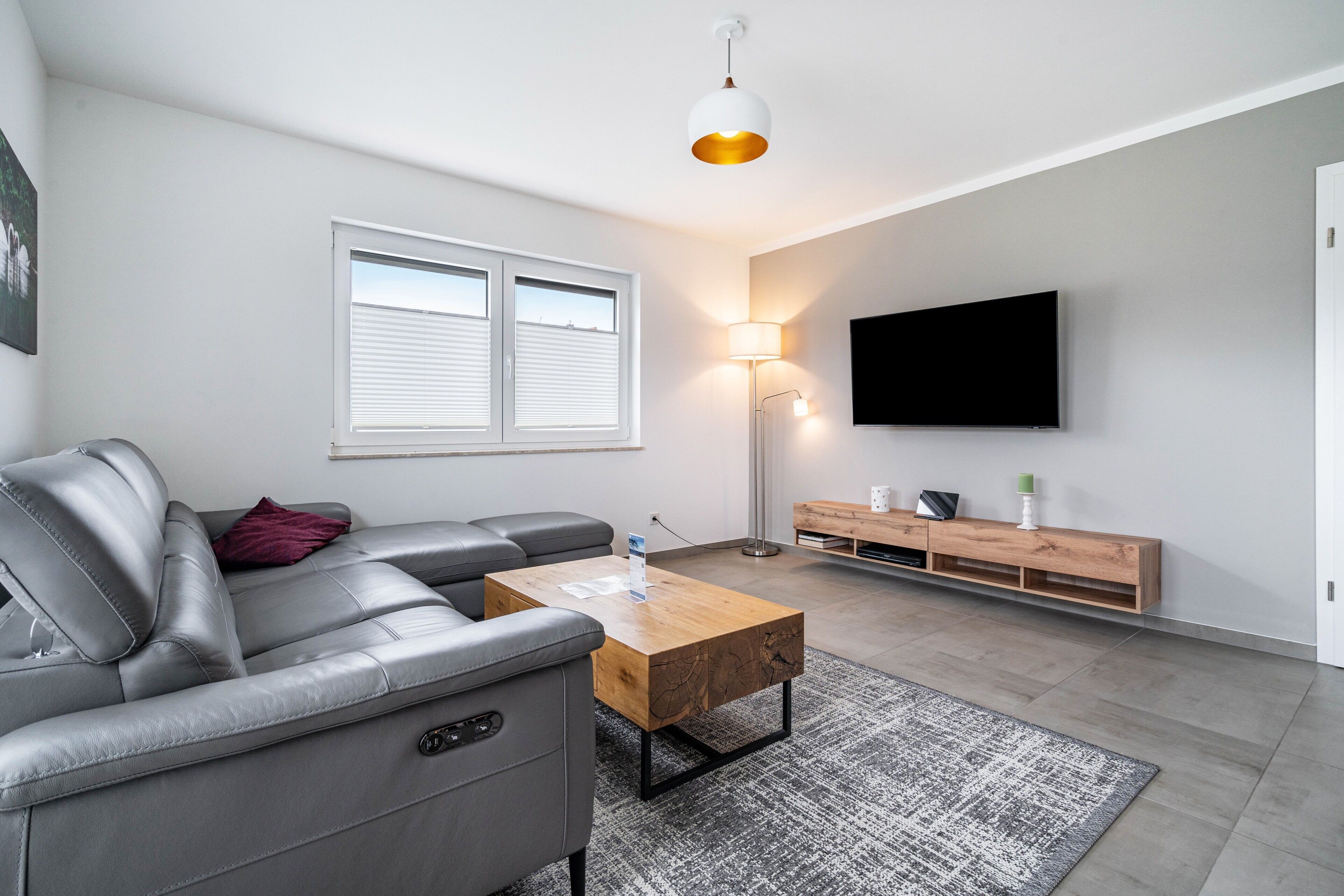 Una sala moderna con un sofá gris, una mesa de centro de madera, una lámpara de pie, un televisor montado en la pared y una ventana con persianas.
