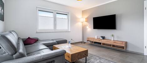 Una sala moderna con un sofá gris, una mesa de centro de madera, una lámpara de pie, un televisor montado en la pared y una ventana con persianas.