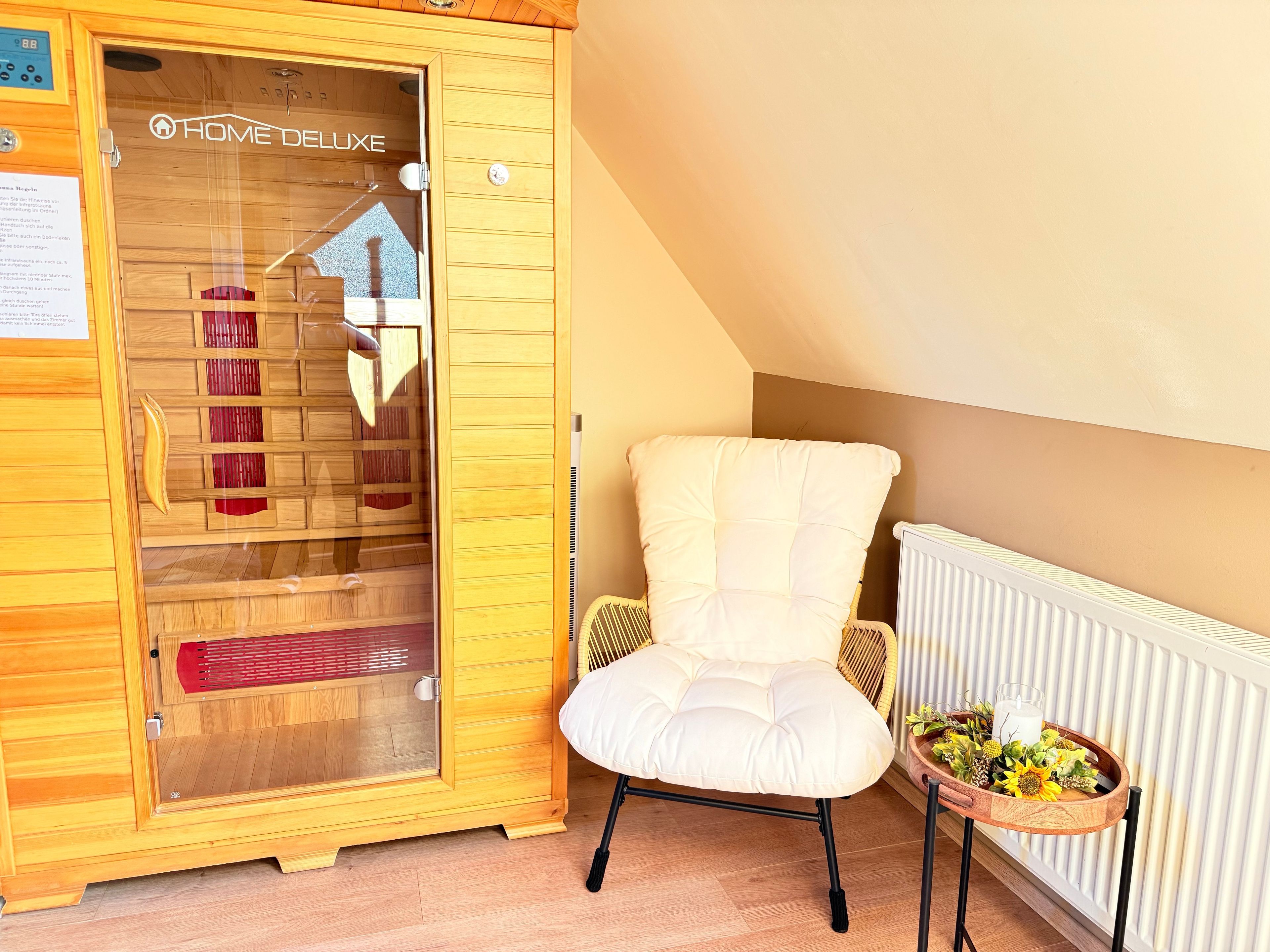 Ein Zimmer mit einer Holzaus, einem weißen gepolsterten Sessel, einem kleinen Tisch mit einer Blumenplatte und einem Glas sowie einer Heizung.