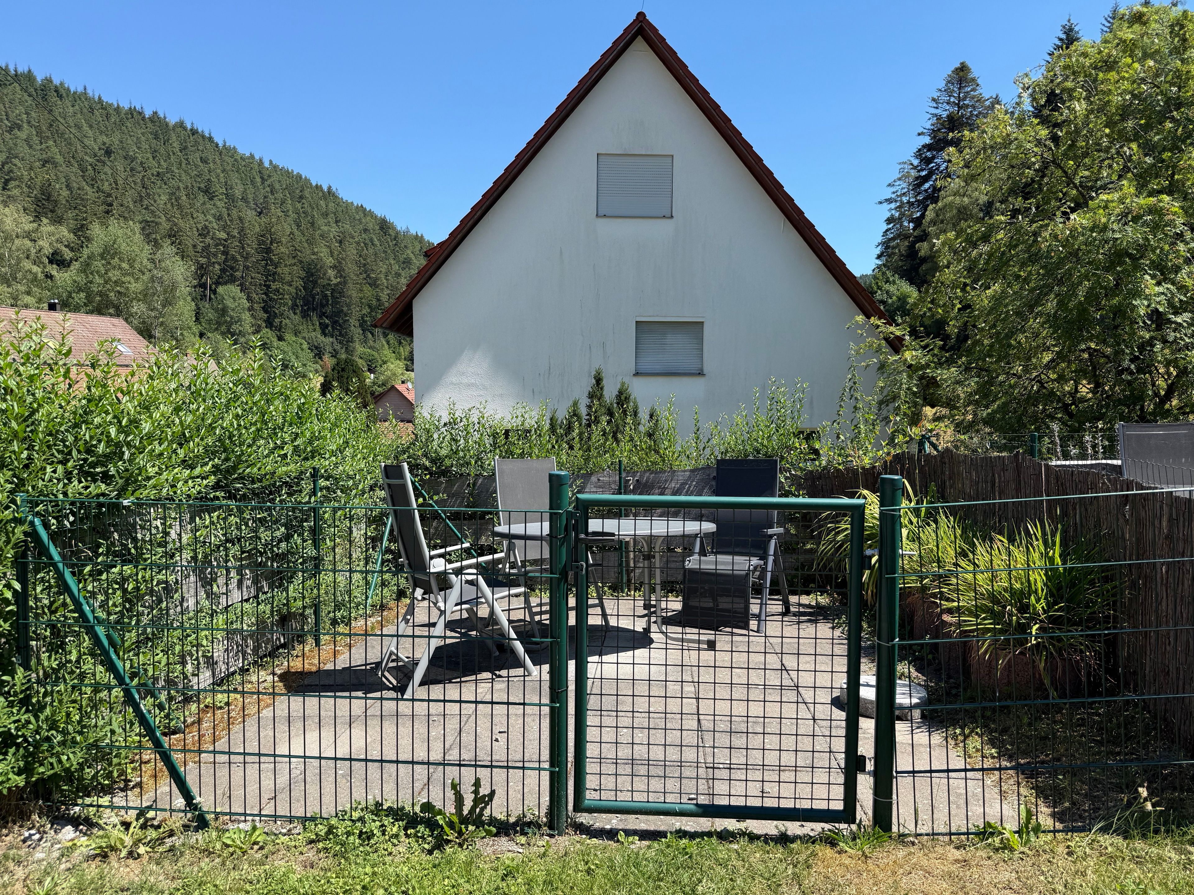 Una casa con patio cercado, mobiliario exterior y vista a una ladera boscosa.