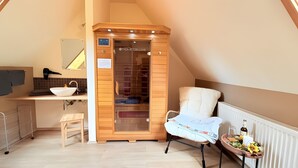 Una habitación pequeña y acogedora con un armario de madera, una silla blanca, una mesita con una botella y flores, y un radiador.