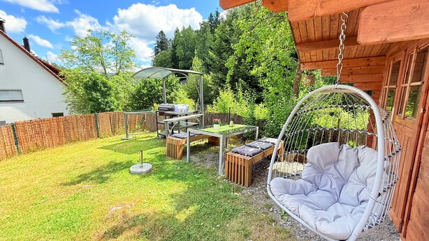 A backyard with a hanging chair, a table with chairs, a grill, and a wooden fence. - Holiday Apartment 'Waldblick' with Fireplace, Fenced Garden, Playground, Grill, Wi-Fi, Sauna (Enzklösterle)