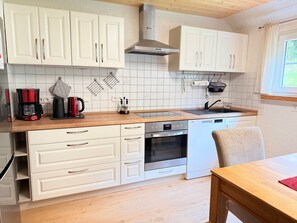 Une cuisine avec des meubles blancs, un plan de travail en bois, une machine à café, une bouilloire, un lave-vaisselle et une fenêtre avec des rideaux.