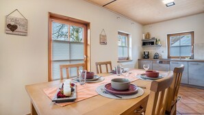 Un comedor con una mesa de madera dispuesta para cuatro, una cocina al fondo y una ventana con persianas.