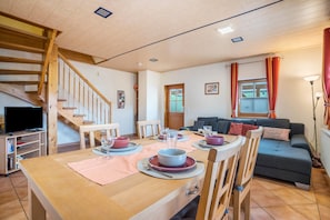 A dining area with a wooden table set for four, a staircase, a living room with a sofa, and a television in the corner.