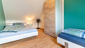 Un dormitorio con dos camas, una mesita de noche con lámpara, un ventilador de pie y un armario.