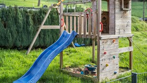 Una casita de juegos de madera con un tobogán y una pared de escalar, rodeada de césped y un seto.