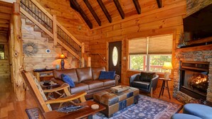 A cozy log cabin interior with a fireplace, a wooden staircase, and a comfortable seating area. - Mountain Cabin, Hot Tub, Fire Pit, Views, Outdoor Bar, Stocked Kitchen (Bryson City)