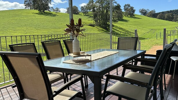 Une terrasse extérieure avec une table et des chaises, un vase rempli de fleurs et une vue sur une colline verdoyante.
