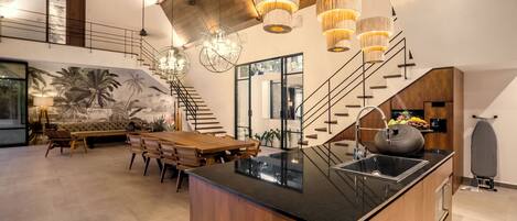 A spacious living area with a wooden dining table, a large island with a sink, and a staircase leading to an upper level.